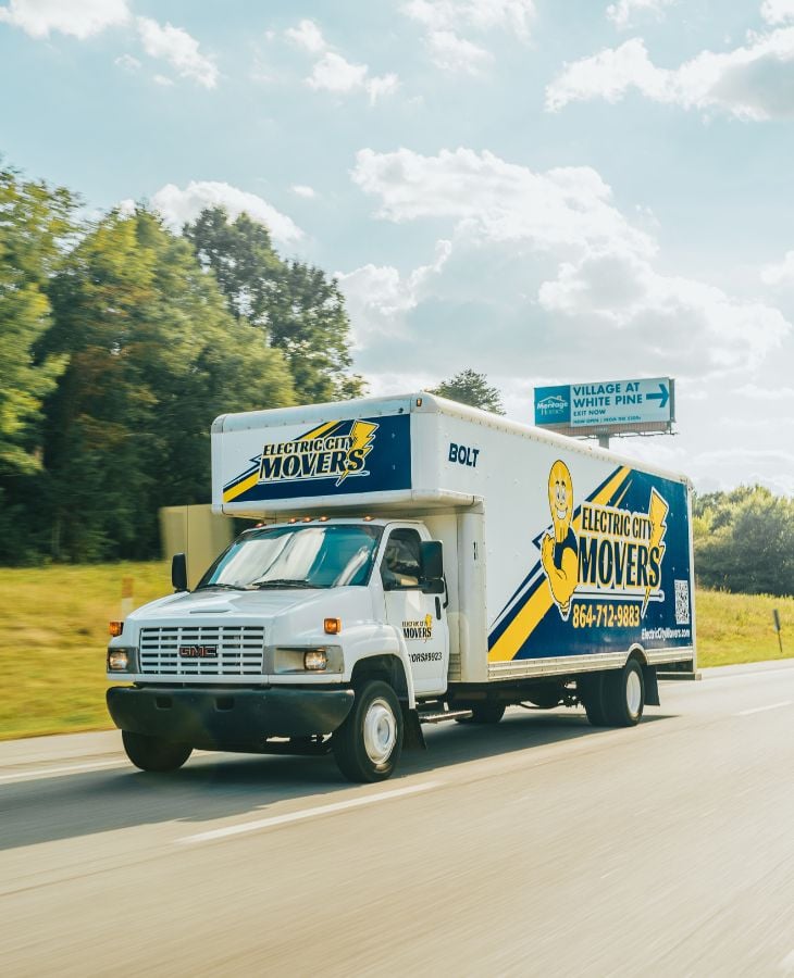 Electric City Movers LLC branded moving truck Bolt driving on a highway surrounded by trees in Anderson, SC, displaying the company logo and phone number 864-712-9883