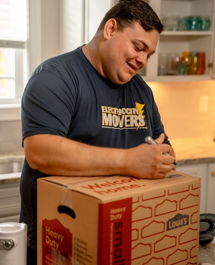Electric City Movers LLC crew member smiling while labeling a moving box in a residential kitchen during a packing and moving service in Anderson, SC