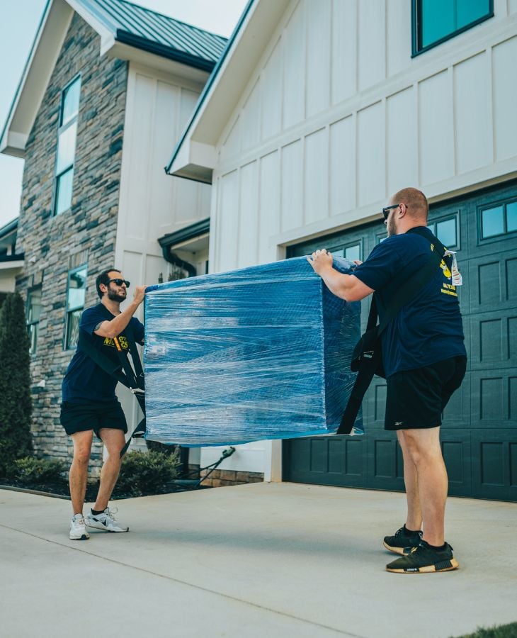 Two Electric City Movers LLC crew members carrying a large plastic-wrapped item in the driveway of a modern stone and board-and-batten residential home during a local move in Anderson, SC
