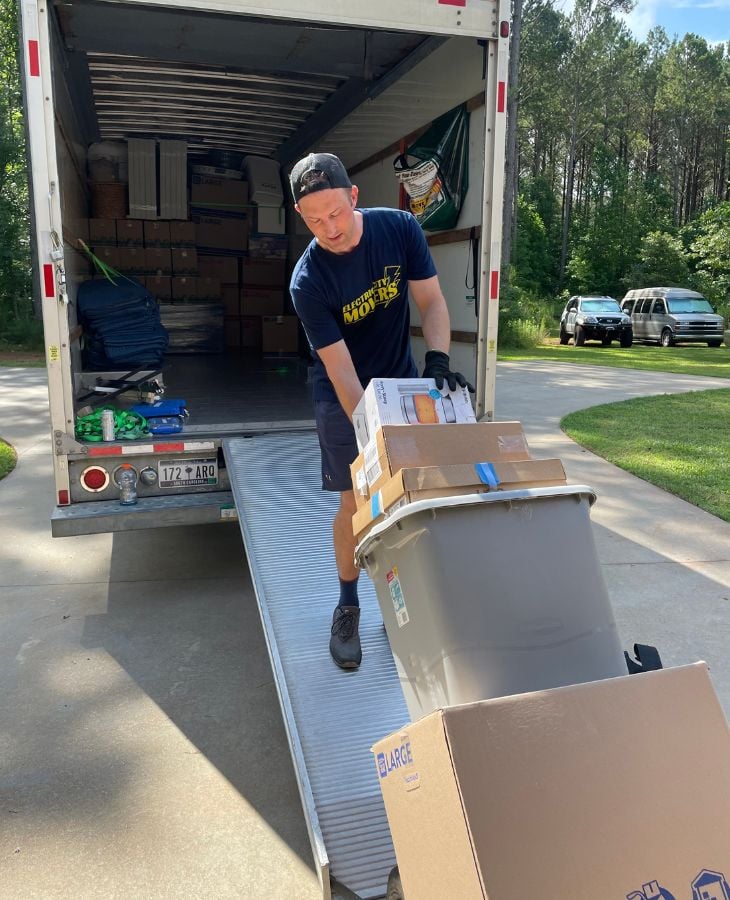 Electric City Movers LLC crew member unloading boxes and belongings from a fully loaded moving truck using a loading ramp at a residential property in Anderson, SC