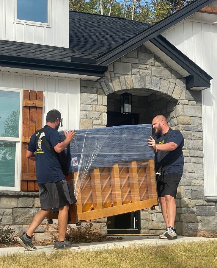 Two Electric City Movers LLC crew members carefully carrying a large plastic-wrapped piece of wooden furniture through the front entrance of a modern stone and wood residential home in Anderson, SC